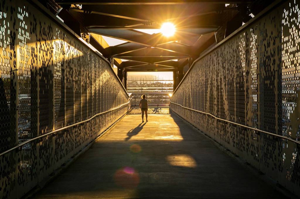 A pedestrian crosses over Marine View Drive toward the waterfront on the Grand Ave Park Bridge as the sun sets Wednesday, in Everett. (Ryan Berry / The Herald)