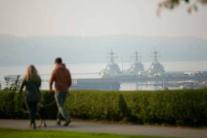 A couple walks their dog through Grand Avenue Park as smoke shrouds Naval Station Everett in the distance on Saturday, Oct. 15, 2022, in Everett, Washington. (Ryan Berry / The Herald)