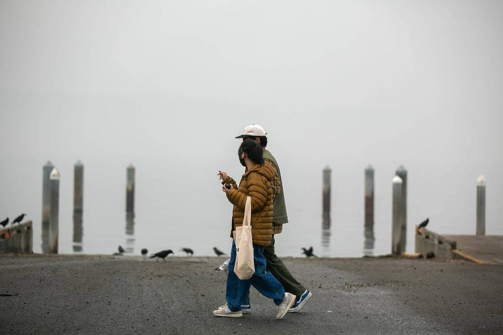 A couple walks along the water at Lighthouse Park, where Whidbey Island would typically be visible in the distance on Thursday, in Mukilteo. Wildfire smoke has been choking the region, but forecasts call for rain in the coming days, which may finally put an end to the haze. (Ryan Berry / The Herald)