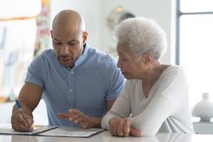 An African American male adviser meets with a female elderly client to discuss her financial plan, life insurance, or medicare needs.  They are both dressed in casual clothing and meeting at a table in the clients home.  It is a very neutral and soft color pallet with whites, grays and blues.  Both the client and the adviser are looking down at the documents on the table.