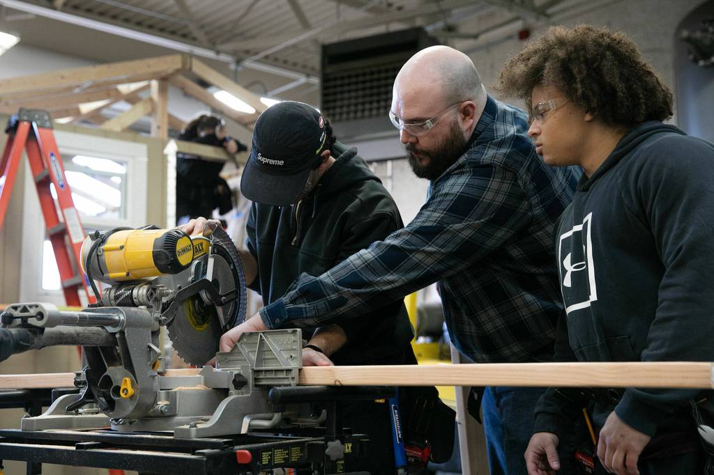 Jeff Lakey, center, and Michael Fields, right, help Darian Vandee as the three work to cut two-by-fours down to size Monday, Dec. 12, 2022, at the Washington Aerospace Training & Research Center in Everett, Washington. (Ryan Berry / The Herald)