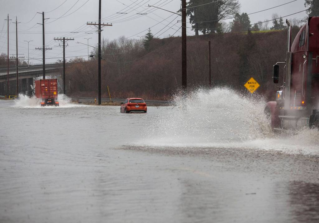 Semi trucks drive around a flooded car along Marine View Drive on Tuesday, Dec. 27, 2022 in Everett, Washington. (Olivia Vanni / The Herald)