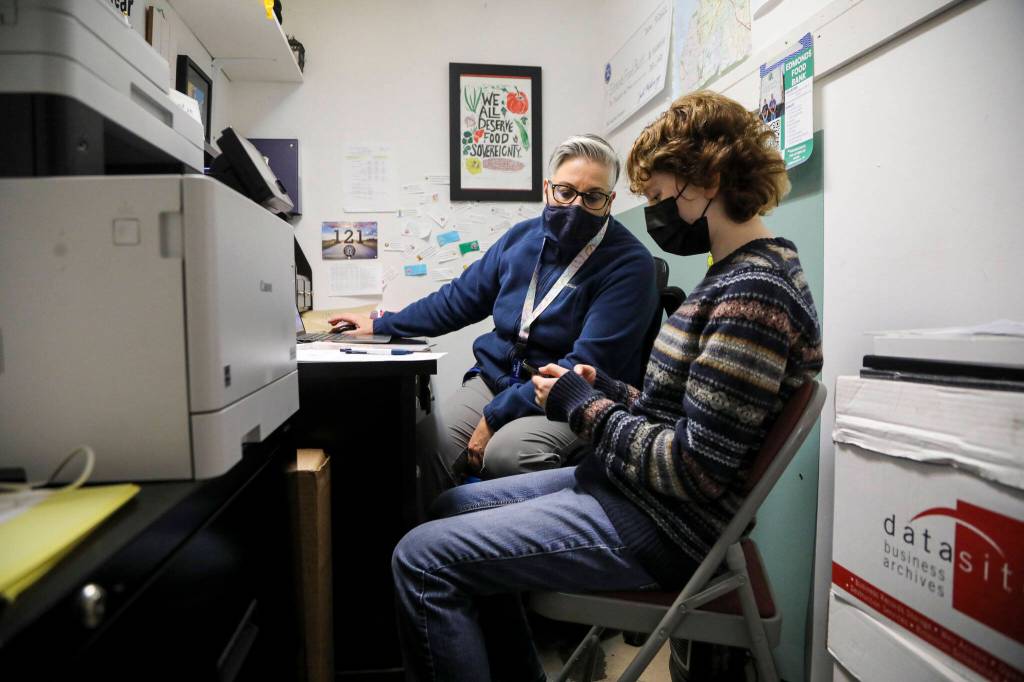 Juniper DeCasso, 17, right, talks through writing a survey with Executive Director Casey Davis at the Edmonds Food Bank in Edmonds, on Jan. 16. Juniper, a Scriber Lake High School student, works at the food bank as part of an on-the-job training class that teaches students about career options and goal planning, while also paying them for a part-time internship. (Annie Barker / The Herald)