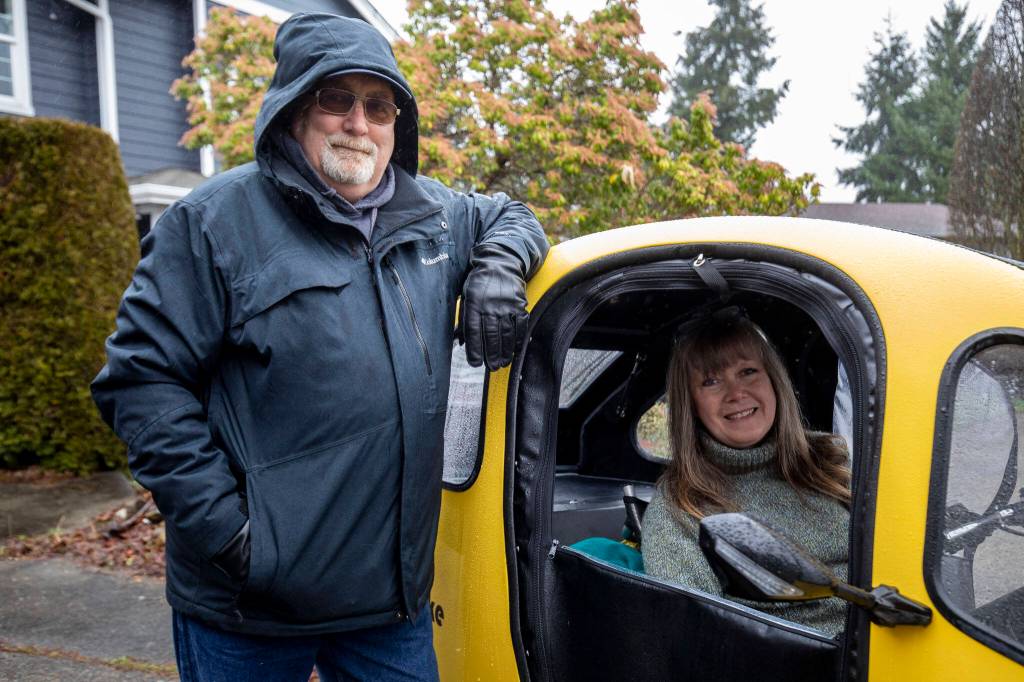 Chris Glans, 65, left, and his wife, Paula Townsell, 62, pose for a photo with their PEBL at their home in Everett. The yellow microcar is an electric bike called PEBL with doors, windows, windshield wiper, lights, trunk and more. (Annie Barker / The Herald)
