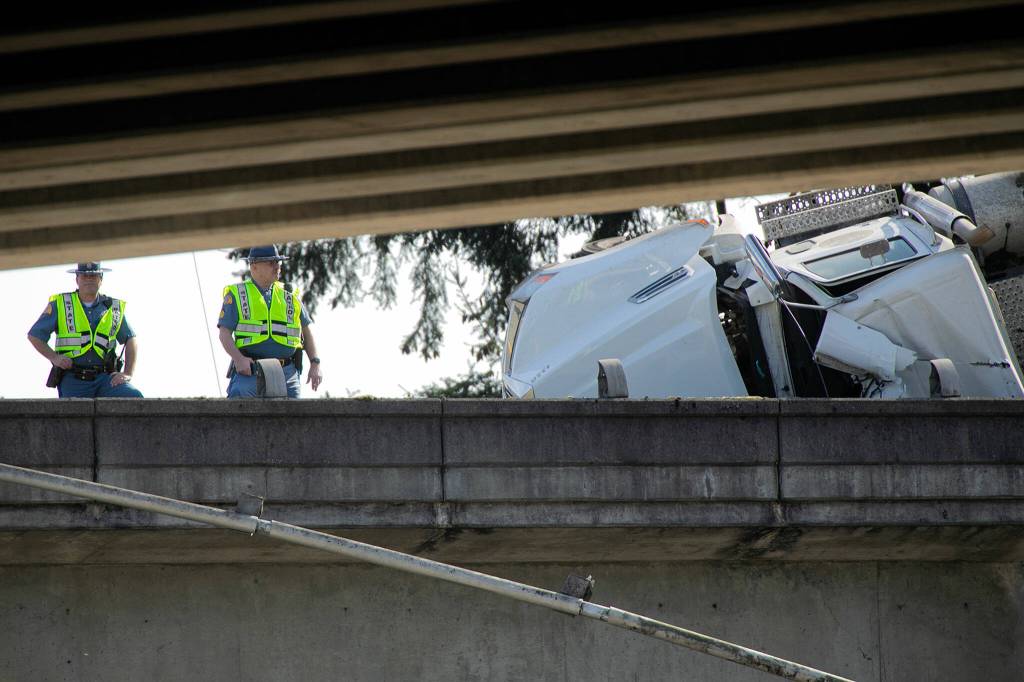 Two state troopers stand near a semi carrying trash as it leans over the edge of I-5 northbound after a late morning crash above Everett Avenue on Thursday, March 16, 2023, in Everett, Washington. (Ryan Berry / The Herald)