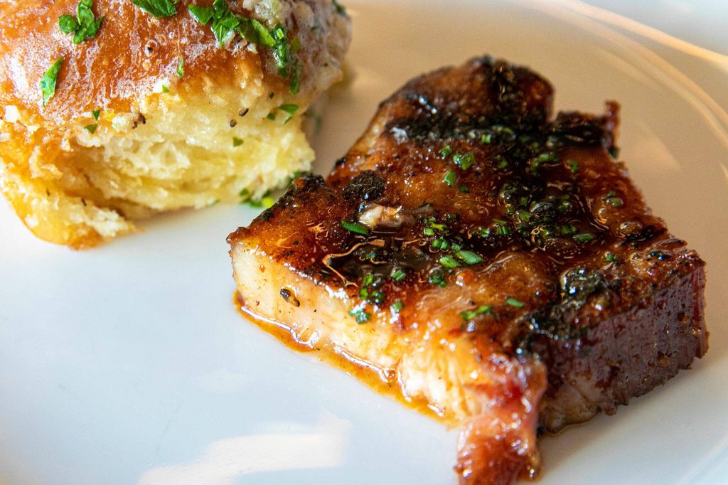 A slice of the most popular appetizer  Bacon in gochujang glaze  is plated alongside a roll at Rare Society on Tuesday, April 25, 2023, in Mill Creek, Washington. (Ryan Berry / The Herald)