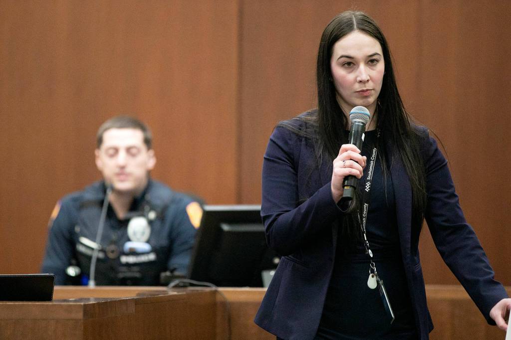 Sarah Johnson walks past the jury while speaking with Everett Police officer Brian Bratlien, background, during the first day of the trial of Shayne Baker on Monday, April 24, 2023, at Snohomish County Superior Court in Everett, Washington. (Ryan Berry / The Herald)