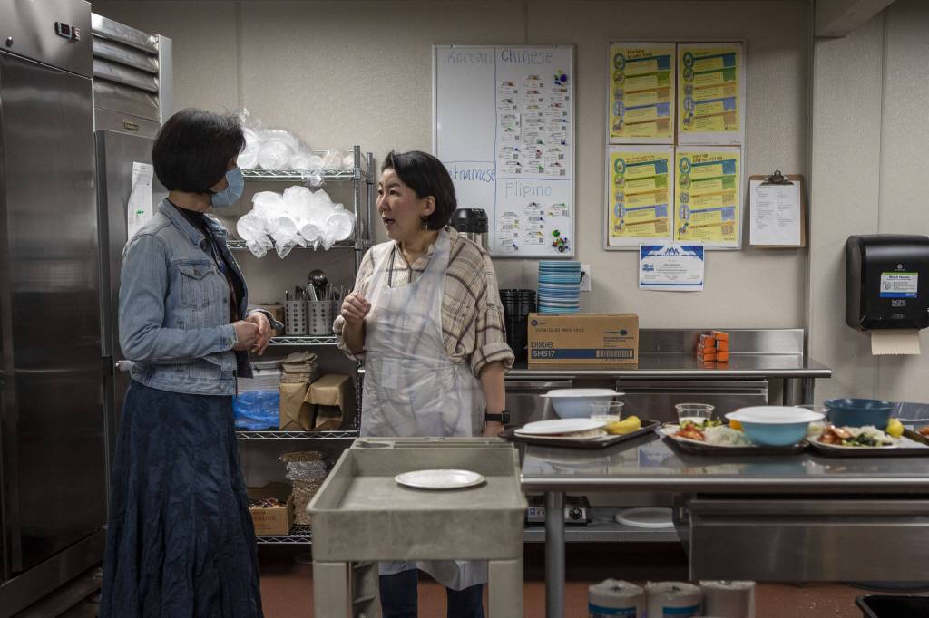 Julie Cho, left, and Jeannie Choi, right, speak in the kitchen during a senior community meals program focused on the Korean community at Homage in Lynnwood, Washington on Thursday, May 4, 2023. (Annie Barker / The Herald)