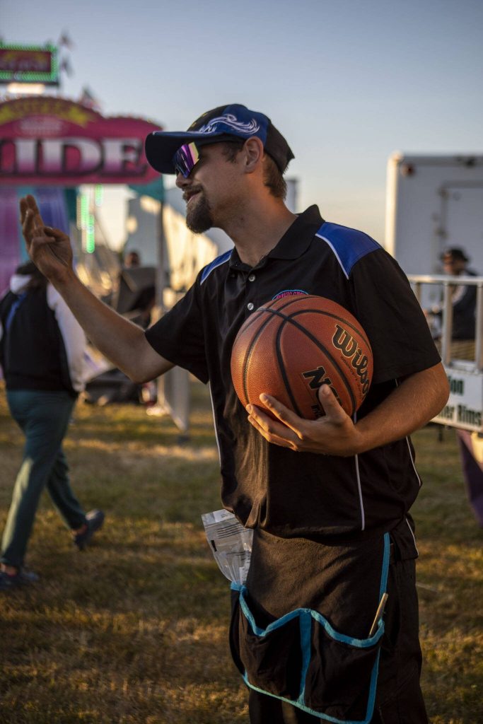 David Crabtree, 20, beckons to people to try shooting a basketball for a prize during the Kla Ha Ya Days carnival at Harvey Field in Snohomish, Washington on Thursday, July 13, 2023. This is Crabtrees first year with the carnival. (Annie Barker / The Herald)