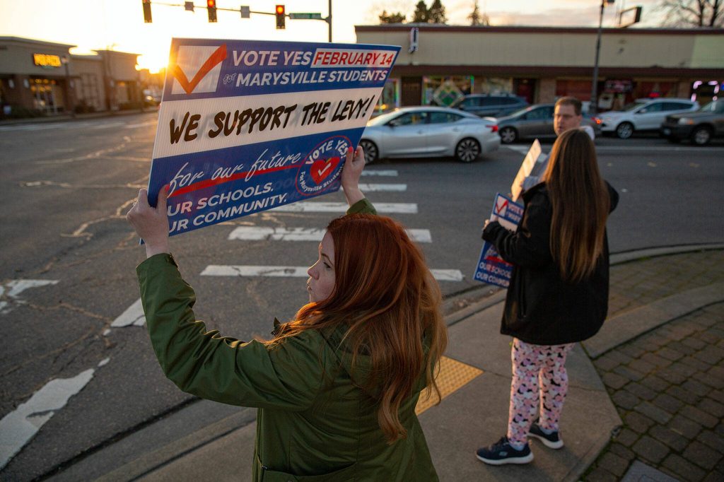 Community advocate and former state representative Emily Wicks waves signs at the corner of 4th and State along with Christie Ryba-Johnson and Matt Ruskowski, both employees at Marysville School District, in support of a school levy in February. (Ryan Berry / The Herald)