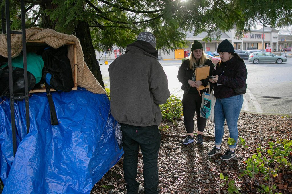 Liz Skinner, right, and Emma Titterness, both from Domestic Violence Services of Snohomish County, speak with a man near the Silver Lake Safeway while conducting a point-in-time count Tuesday, Jan. 23, 2024, in Everett, Washington. The man, who had slept at that location the previous night, was provided some food and a warming kit after participating in the PIT survey. (Ryan Berry / The Herald)