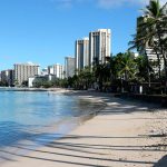 FILE - A man sits on a nearly empty Waikiki Beach in Honolulu, Friday, Oct. 2, 2020. Tourists are traveling to Hawaii in larger numbers than officials anticipated, and many are wandering around Waikiki without masks, despite a statewide mandate to wear them in public. (AP Photo/Caleb Jones, file)