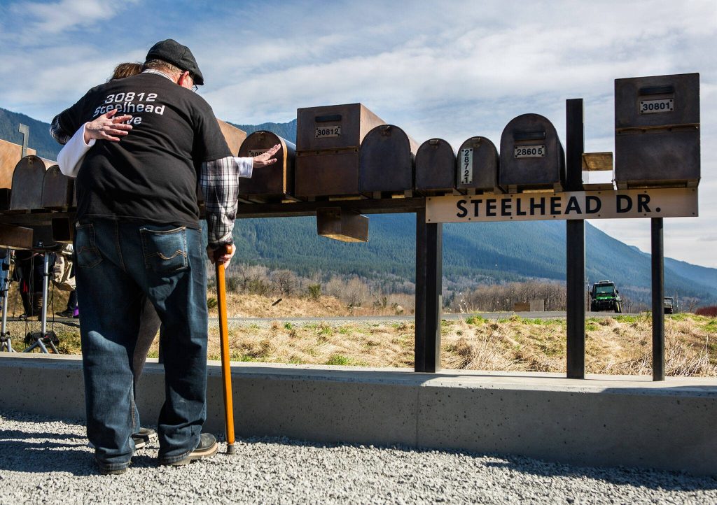 Ron Thompson, right, embraces his wife Gail Thompson, left, as she reaches out to touch a replica of their mailbox during the Oso slide remembrance ceremony on Friday, March 22, 2019 in Oso, Washington. (Olivia Vanni / The Herald)