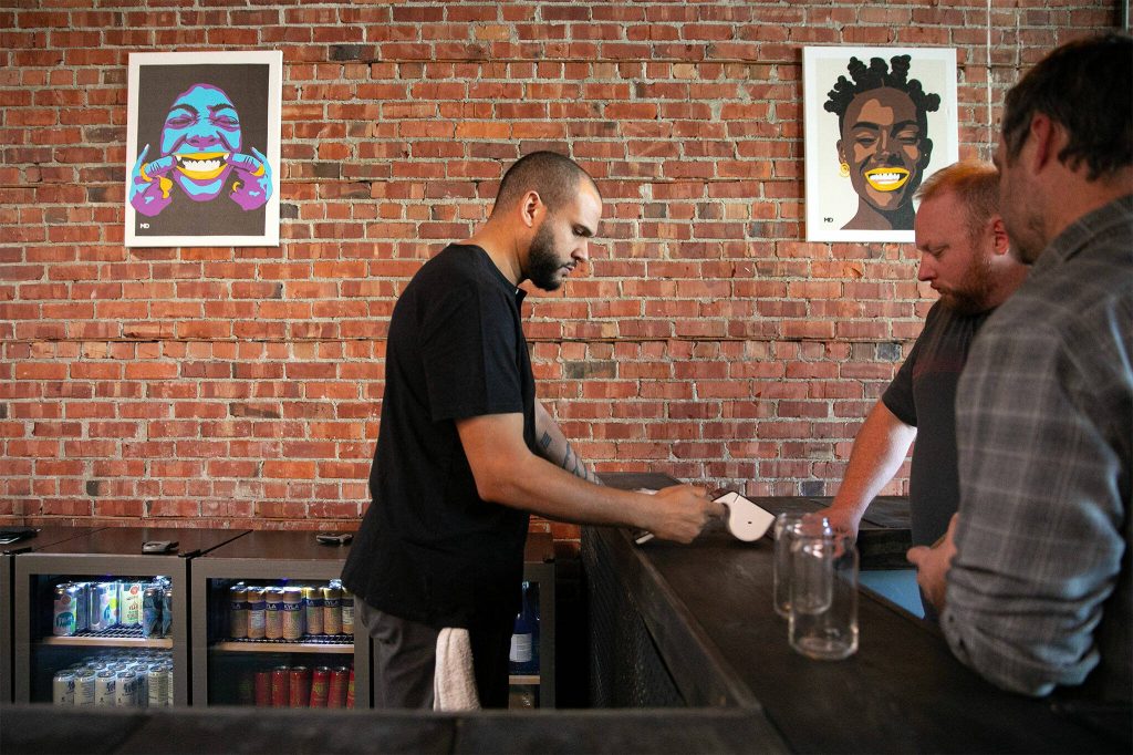 Craig Chambers takes orders while working behind the bar at Obsidian Beer Hall on Friday, April 12, 2024, in downtown Everett, Washington. Art by Jessie Lipscomb or Momma Lips Draws hangs behind him. (Ryan Berry / The Herald)