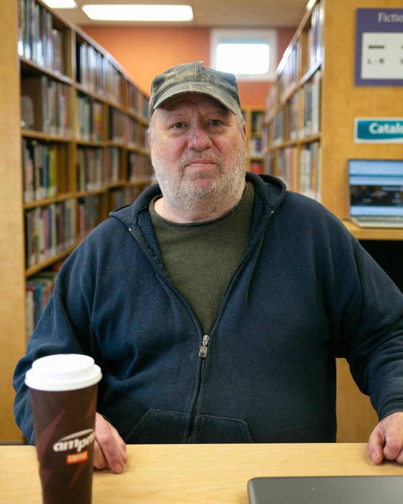 George Beard takes a seat at his spot inside the Stanwood Library on Thursday, May 23, 2024, in Stanwood, Washington. The space is free to use and enables Beard to search for housing online and keep up on his paperwork as he seeks shelter and financial support. (Ryan Berry / The Herald)