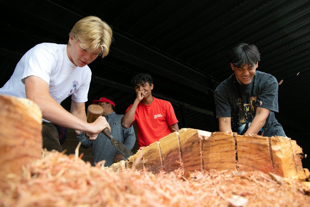 Archbishop Murphy students, from left, Logan Kendrick, Solomon Perera and Noah Jimenez put their backs into their work while carving away excess wood from a healing pole on Tuesday, June 13, 2023, at their school in Everett, Washington. Twenty or so juniors from Roger Brodniak’s APUSH class gathered to help make progress on the redwood artwork. (Ryan Berry / The Herald)