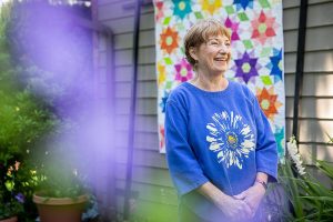 Susan Davison smiles in the shaded section of her yard in front of a quilt titled Dancing Dahlias on Tuesday, July 2, 2024 in Mukilteo, Washington. (Olivia Vanni / The Herald)