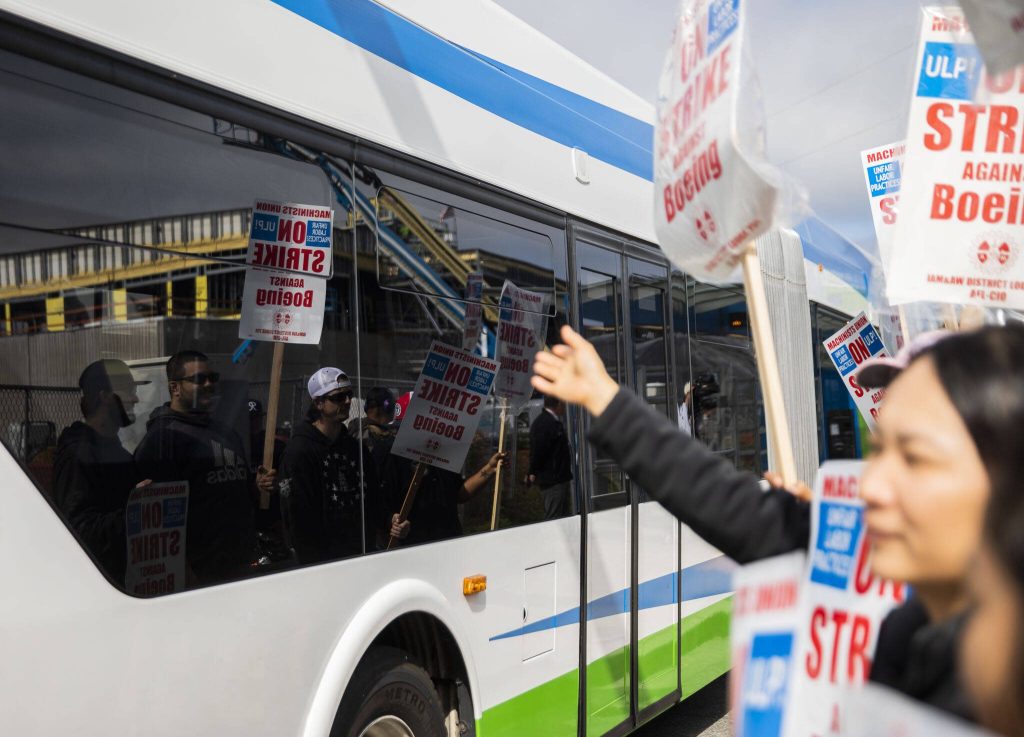 Striking Boeing workers are reflected in the windows of a bus as it drives along Airport Road on Monday, Sept. 16, 2024, in Everett, Washington. (Olivia Vanni / The Herald)