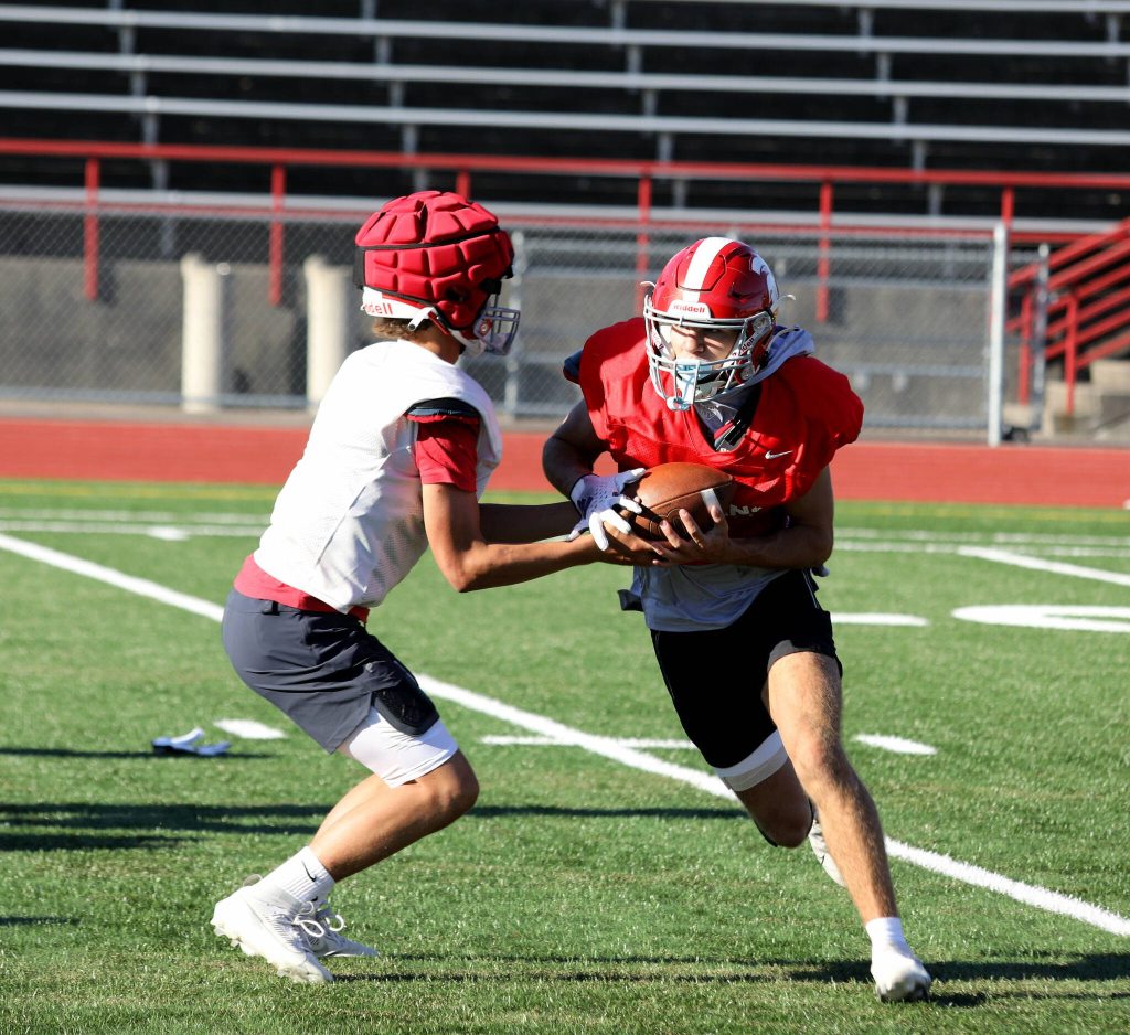 Sophomore quarterback Alex Maldonado (left) hands off the football to senior safety and running back Canyon Bumgarner during the Stanwood High School football practice at Bob Larson Stadium in Stanwood, Washington on Aug. 30, 2024. Some players wore Guardian Caps during practice. (Taras McCurdie / The Herald)