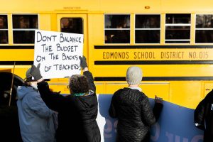 Melody Schneider holds a sign protesting pay cuts to teachers as an Edmonds School District bus passes by during Edmonds College faculty union rally as part of a national day of action outside of the Lynnwood Event Center on Tuesday, March 4, 2025 in Lynnwood, Washington. (Olivia Vanni / The Herald)
