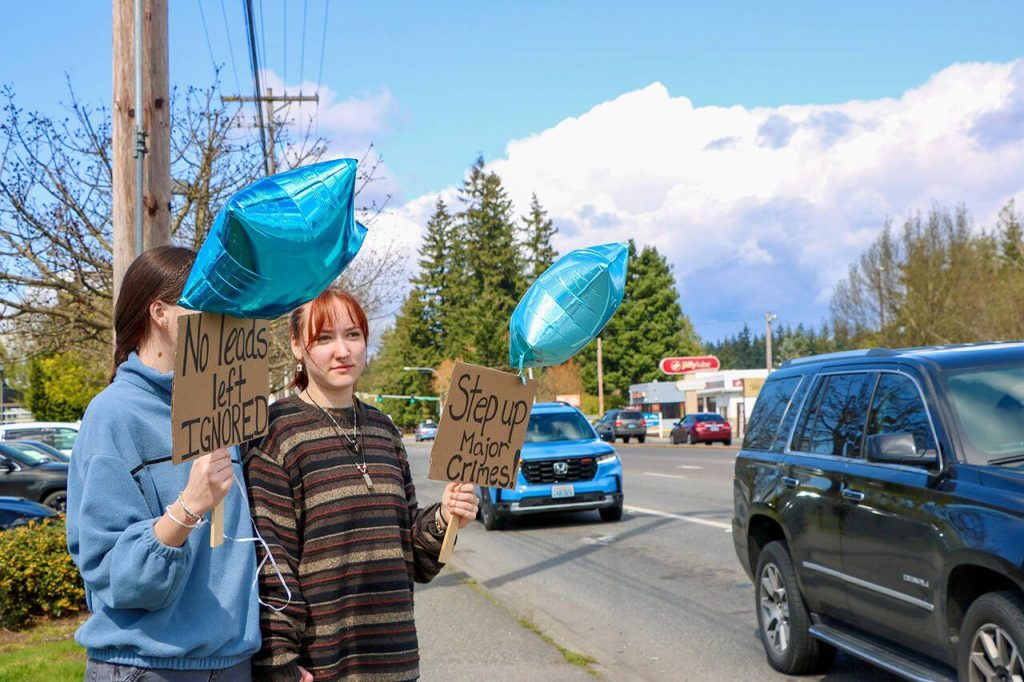 Two protesters hold blue balloons and signs reading No Leads Left Ignored and Step Up Major Crimes during a call to action Saturday, April 12, 2025, in Arlington, Washington. (Aspen Anderson / The Herald)