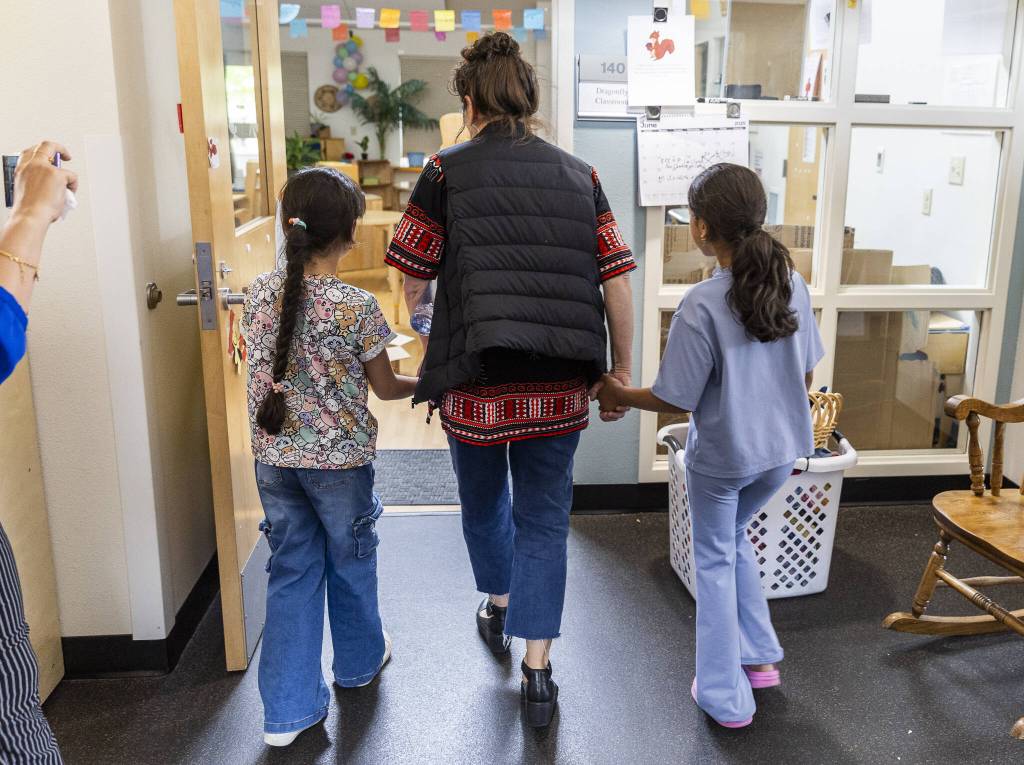 Preschool teacher Michelle Depew walks hand in hand with former Early Learning Center students Lareen Almadjidi, 7, left, and Qamar Almadjidi, 9, right, as they explore their old classroom on Wednesday, June 25, 2025 in Everett, Washington. (Olivia Vanni / The Herald)