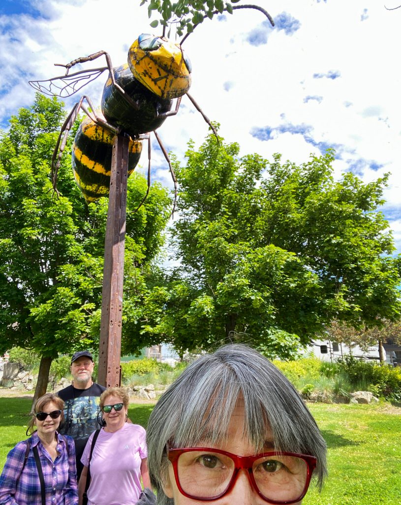 Sue Misao takes a selfie under the yellow jacket sculpture Beest in Twisp with writers Janice Podsada, at left, Andrea Brown, at right, and Max Brown, back. The sculpture looms 15 feet above the Twisp Commons Park and was made mostly from old cars retrieved from the Methow River. (Sue Misao)