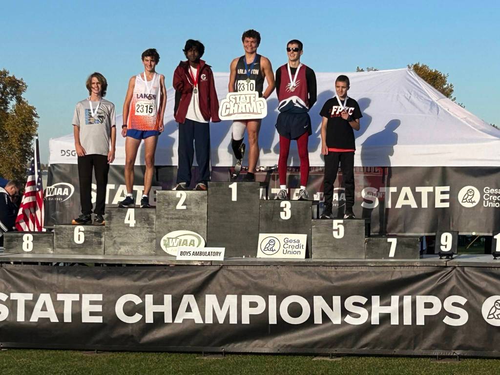 Arlington junior Ramon Little (center) holds the State Champion sign atop the podium after winning the WIAA Cross Country Ambulatory State Championship at Sun Willows Golf Course in Pasco on Nov. 8, 2025. (Photo courtesy Krissy Kolbeck)
