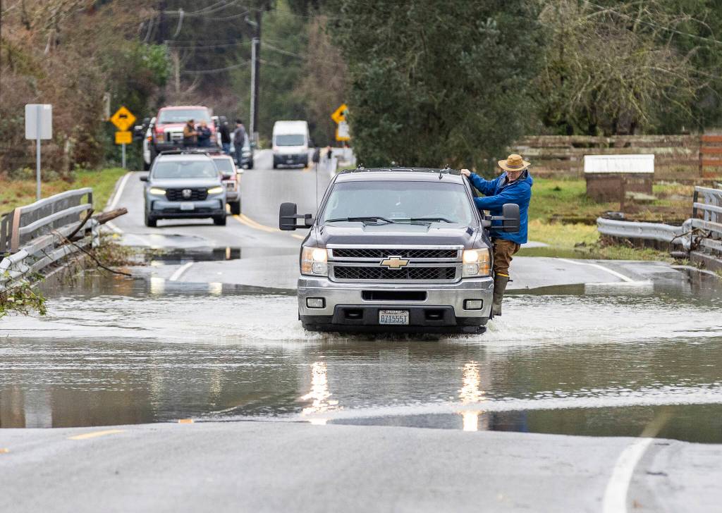 A person hangs off a truck while it drives through floodwater from the Skykomish River covering a portion of Mann Road on Tuesday, Dec. 9, 2025 in Sultan, Washington. (Olivia Vanni / The Herald)