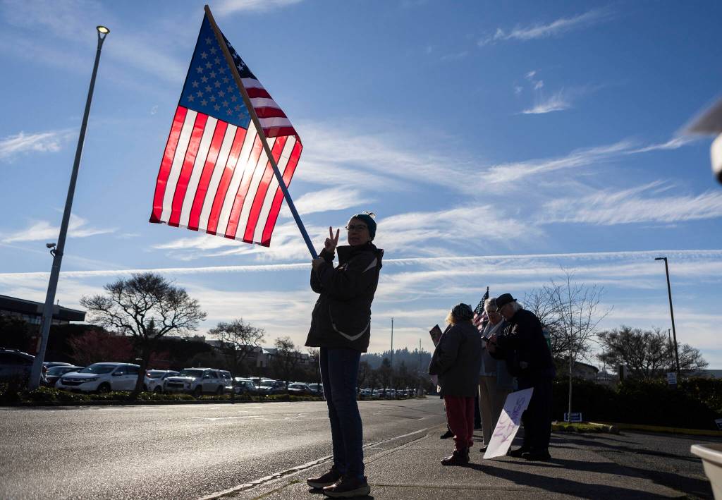 Valerie Heer holds an American flag and flashes a peace sign with her hand to cars driving along Sunset Avenue during South Snohomish County Indivisibles Signs of Fascism protest on Tuesday, Jan. 20, 2026 in Edmonds, Washington. (Olivia Vanni / The Herald)