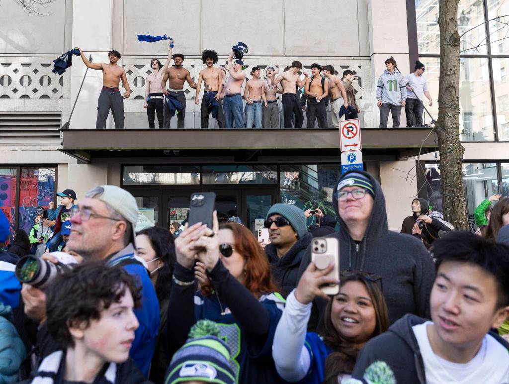 A group climbs up on an awning and waves their shirts in the air during the Seahawks World Champions Parade on Wednesday, Feb. 11, 2026 in Seattle, Washington. (Olivia Vanni / The Herald)