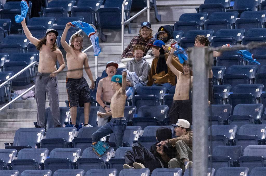A group of kids yell and wave their shirts after the AquaSox score their first run of the game during the fourth inning against the Tri-City Dust Devils on Tuesday, April 7, 2026 in Everett, Washington. (Olivia Vanni / The Herald)