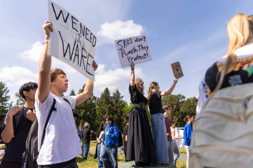 Stefan Krzeszowski, left to right, Quinn Hall and Olivia Santoro hold signs along Bothell Way in protest of the school boards vote to not renew the contract for Bothell High Schools school resource officer on Monday, May 18, 2026, in Bothell, Washington. (Olivia Vanni / The Herald)
